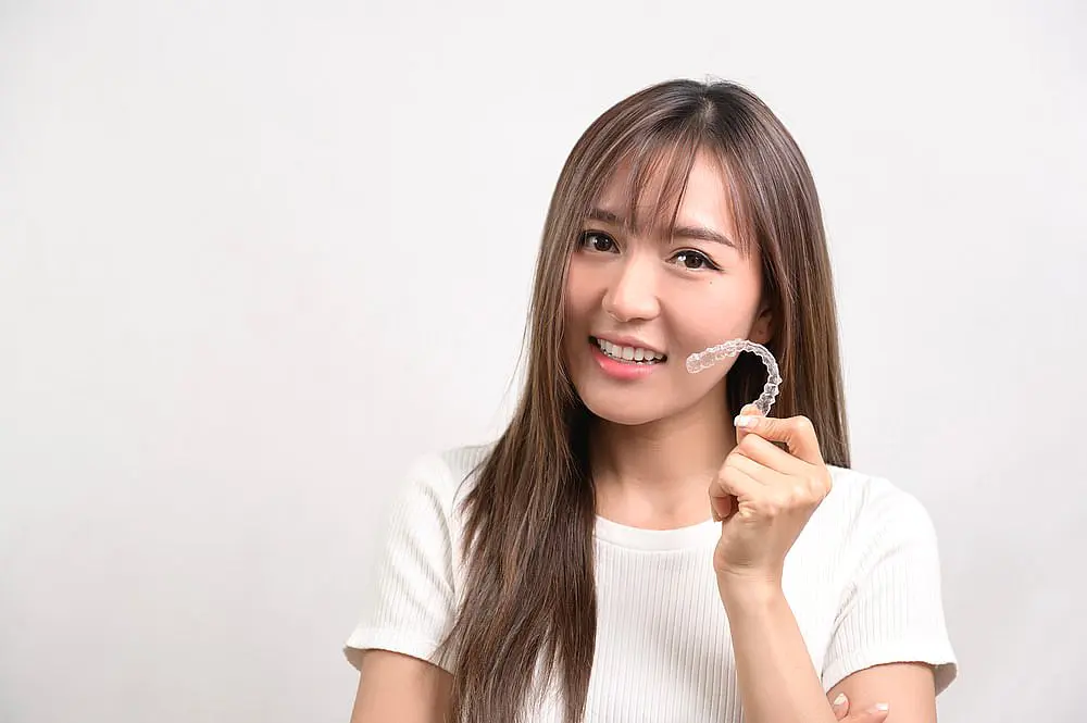 A person with long hair smiles confidently while holding an Invisalign clear dental aligner against a plain background in Tucker, GA