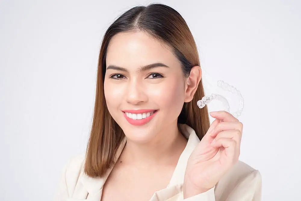 Woman with shoulder-length hair smiling and holding a clear Invisalign aligner, showcasing her stunning smile, all while enjoying the peace of mind provided by her comprehensive dental insurance against a plain background in Suwanee or Tucker, GA