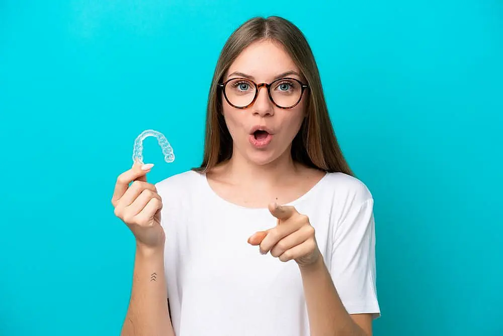 A woman with glasses holds a clear Invisalign dental retainer and points forward against a turquoise background, wearing a white shirt, highlighting the importance of insurance coverage for your smile in Suwanee or Tucker, GA