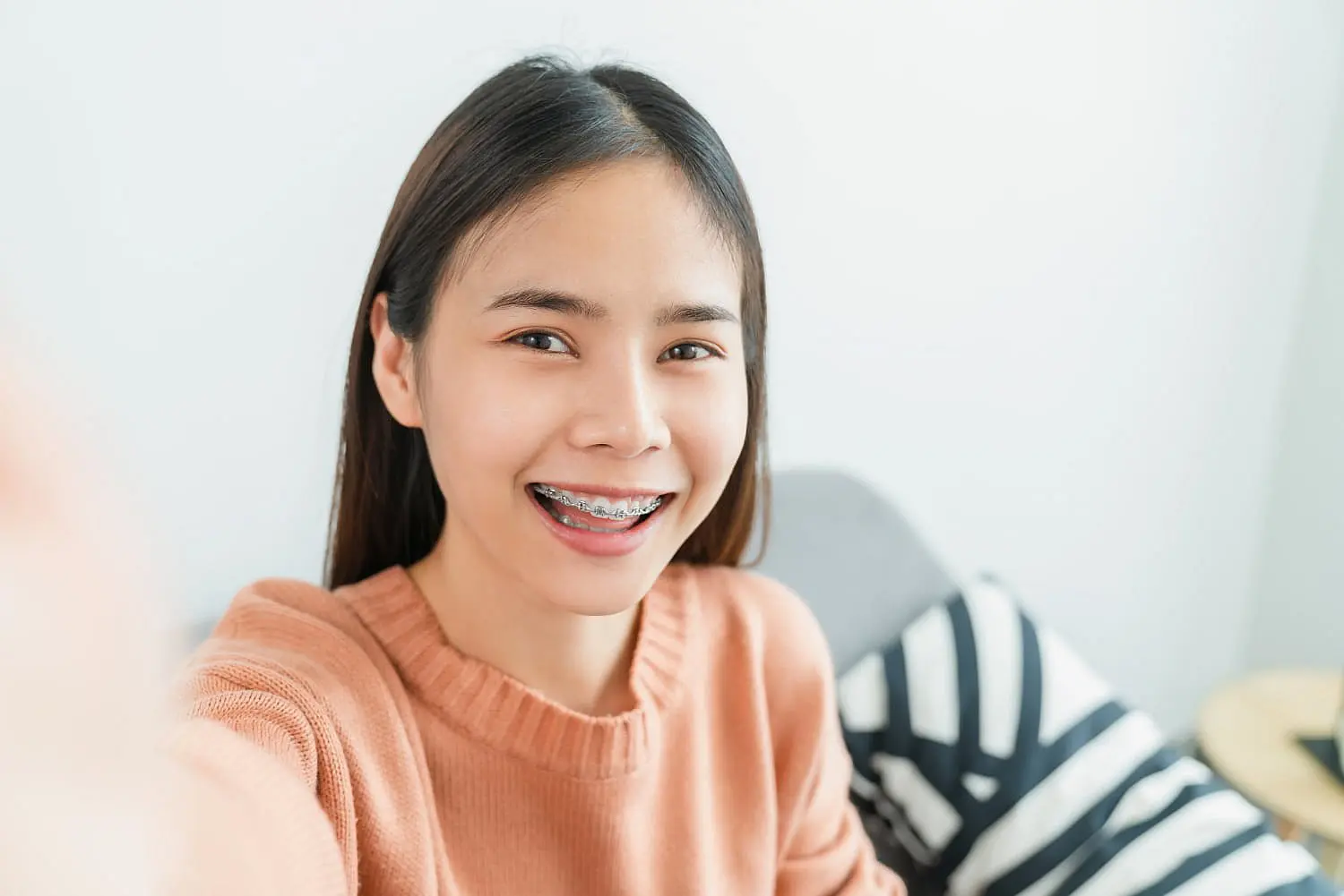 A woman with long hair and braces beams at the camera, her peach sweater complementing her smile. She's seated on a couch adorned with black and white pillows, reflecting the exceptional care she received from NK Orthodontics in Tucker, GA