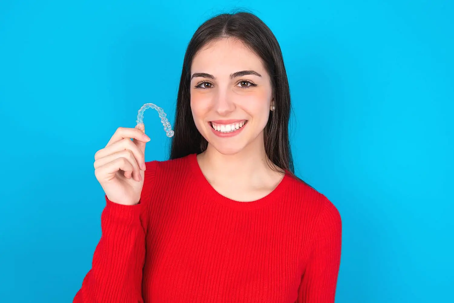 A person in a red sweater holds a clear Invisalign aligner, smiling brightly against a vivid blue background in Suwanee, GA