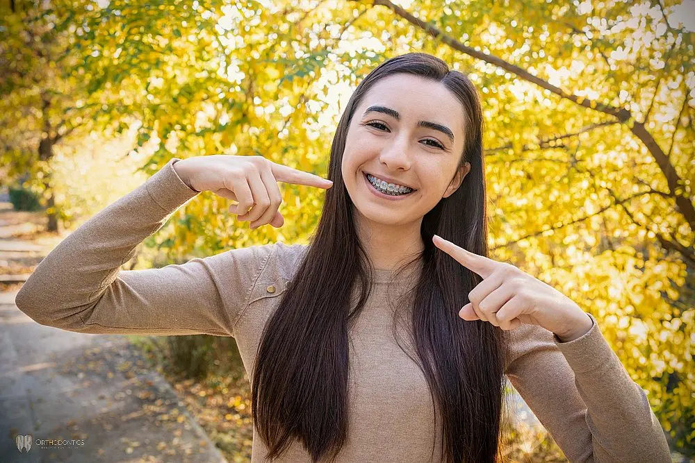 A woman with long hair and braces is smiling and pointing at her braces with both hands after considering Aligners vs Braces in Suwanee & Tucker, GA. She is standing outdoors in front of trees with yellow leaves.