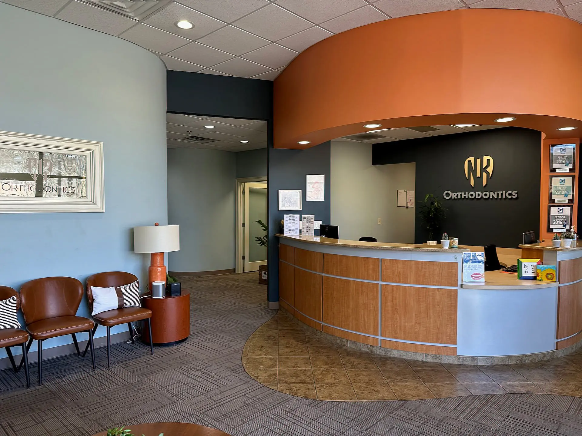 NK Orthodontics features a curved reception desk, vibrant orange accent wall, and cozy seating area, all complemented by proudly displayed certificates in Suwanee, GA