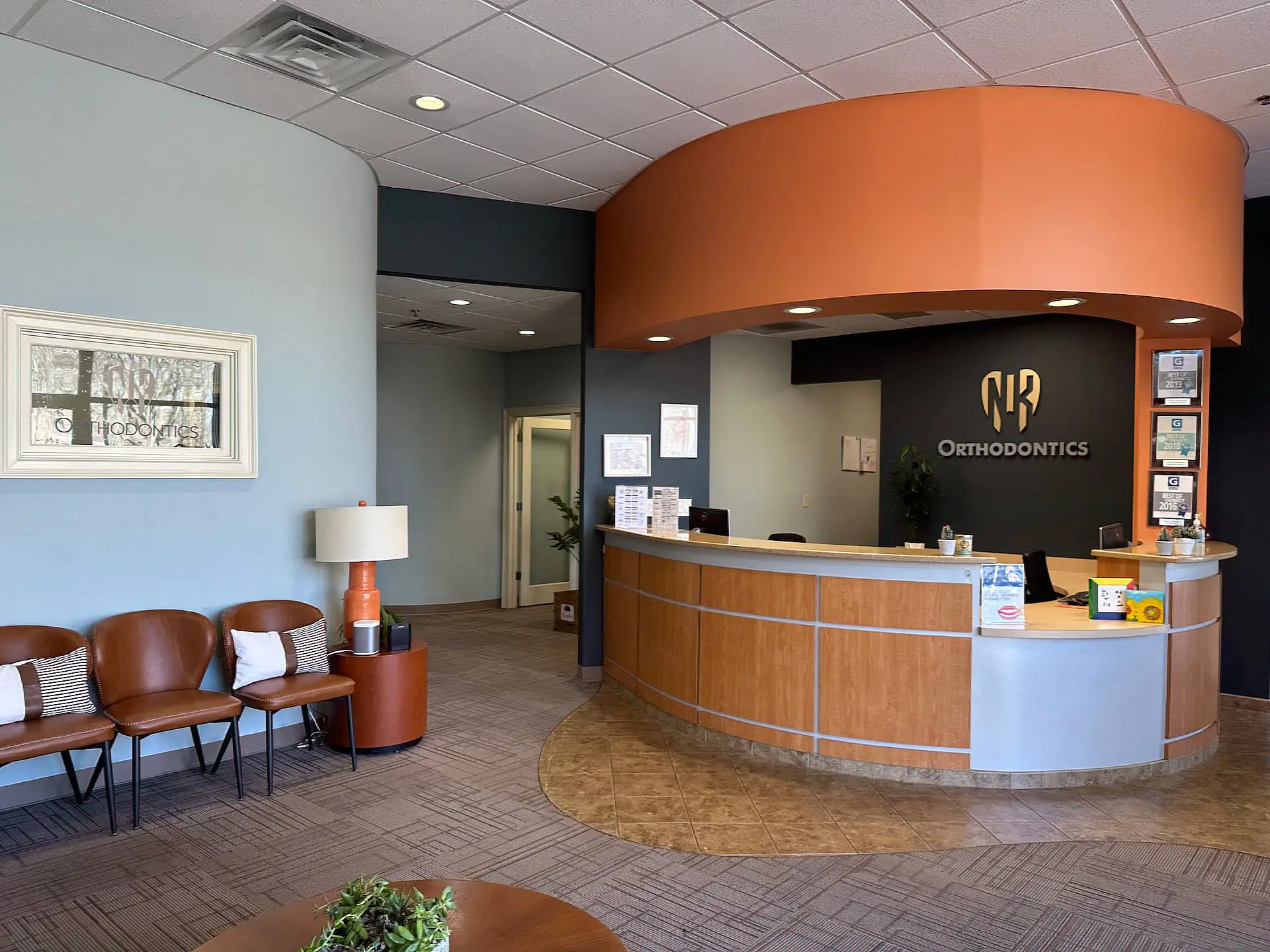 The NK Orthodontics Suwanee reception area boasts a curved desk, vibrant orange and gray walls, comfortable chairs, a lush potted plant, and elegant framed art in Suwanee or Tucker, GA