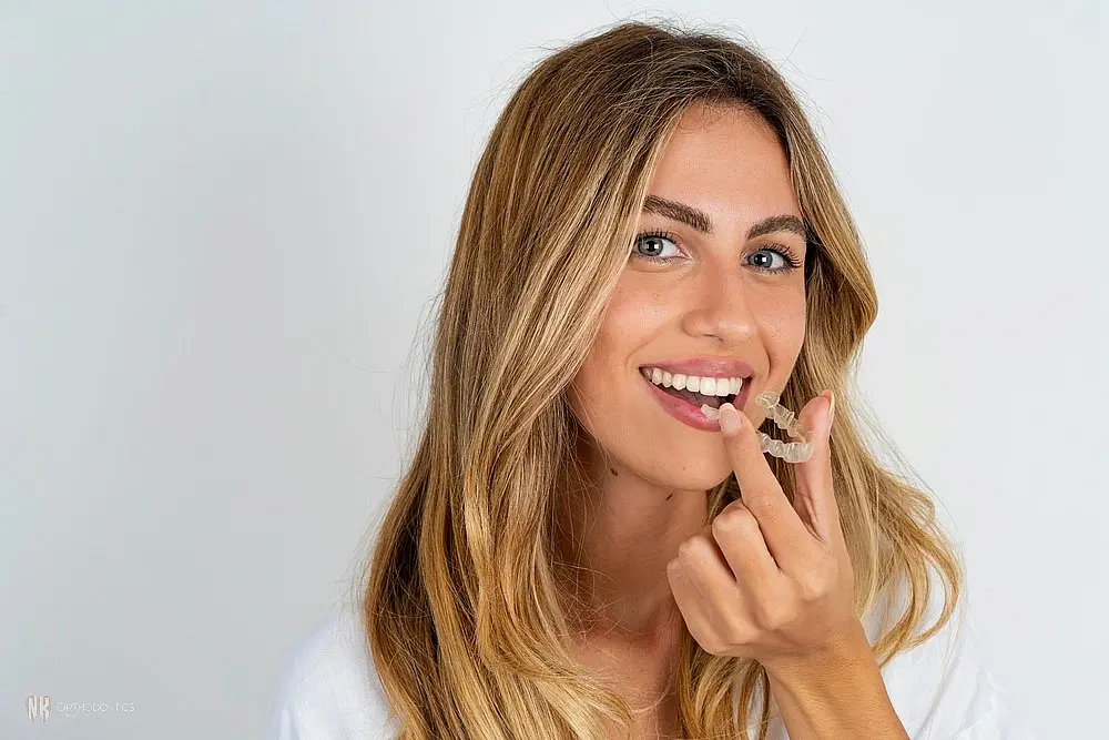 A woman with long hair is holding a clear aligner near her mouth while smiling after considering Aligners vs Braces in Suwanee & Tucker, GA against a plain background.