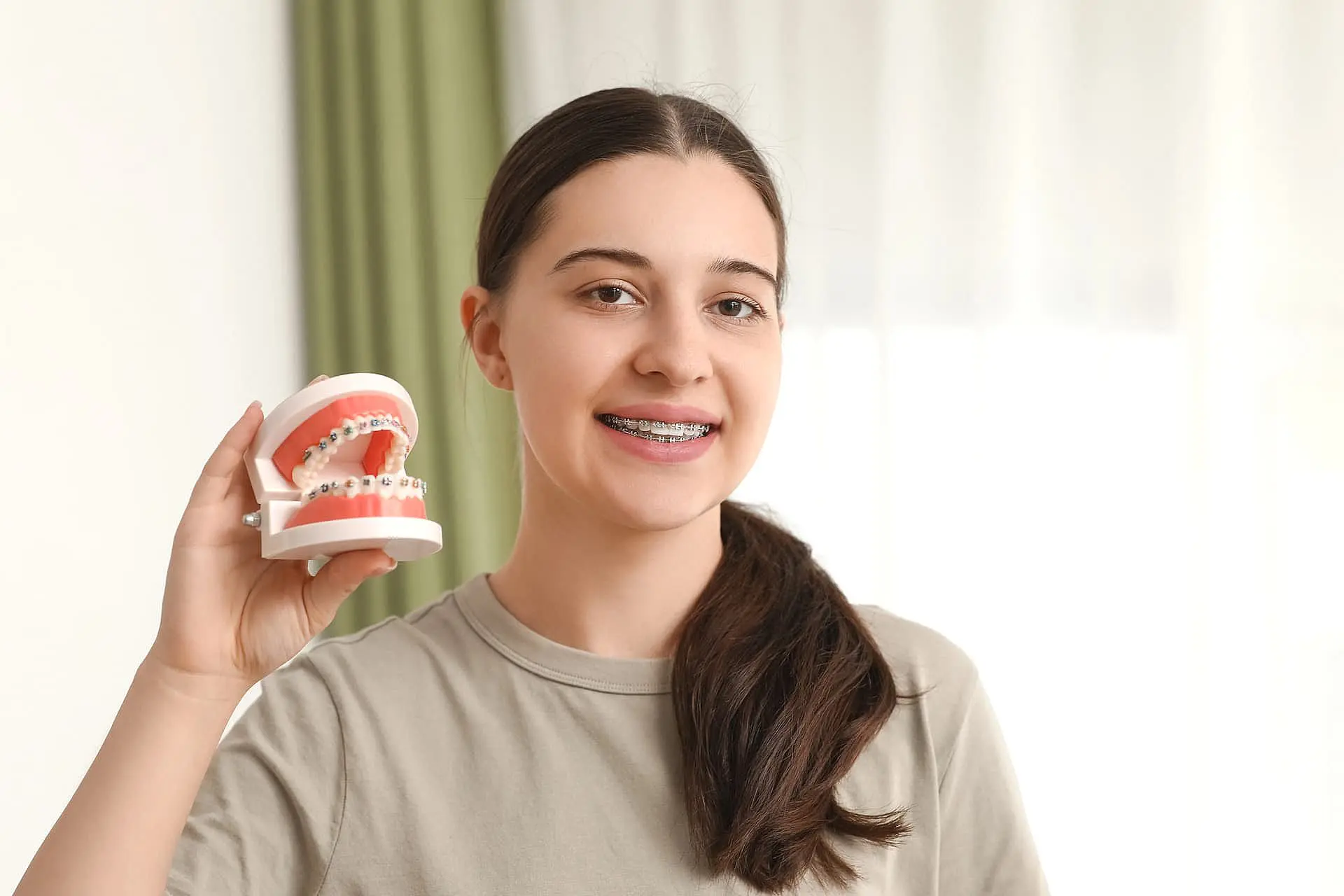 A person with affordable braces holds a dental model showcasing teeth with braces, standing in front of a light-colored curtain in Suwanee or Tucker, GA