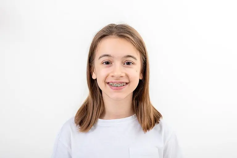 A young person with straight, shoulder-length brown hair and braces smiles at the camera against a plain white background, ready to learn How Long Do Braces Take in Suwanee and Tucker, GA.