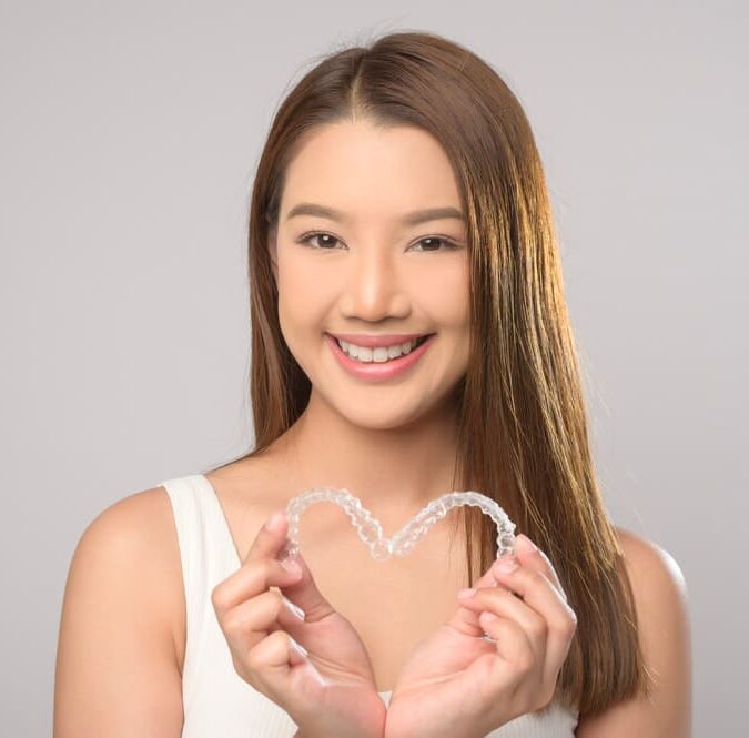 A woman with long hair beams as she holds an Invisalign aligner shaped like a heart against a plain background, showcasing the joy of adult orthodontics in Suwanee or Tucker, GA