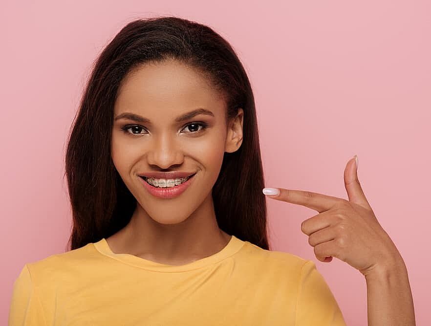 A person in a yellow shirt joyfully points to their clear braces, set against a vibrant pink background in Suwanee, GA